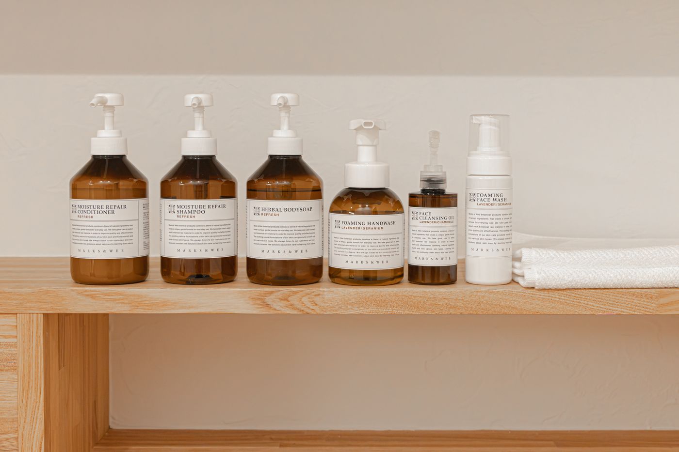 Bath amenities lined up on a wooden shelf: shampoo, conditioner, body soap, hand wash, cleansing oil and face wash, with a folded towel